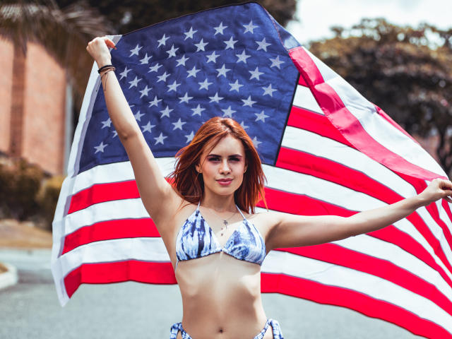 AlmaBella with red hair, holding a large American flag, wearing a blue and white tie-dye bikini, arms raised, showing her tits, standing outdoors, smiling confidently.