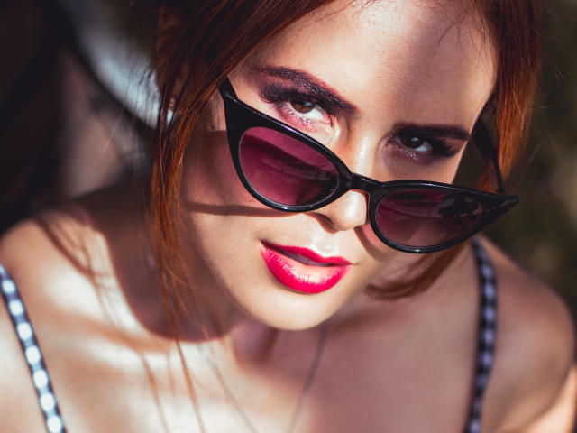 AlmaBella has shiny red hair, black cat-eye sunglasses, pink glossy lips, and is wearing a black-and-white polka dot top, looking up with a slight smile.