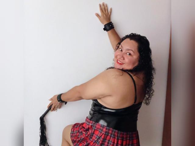 Curvy HiyunaMei with black curly hair, red plaid skirt, black leather top, and wrist cuffs, smiling, hand on wall, holding black leather whip, tattoo on right shoulder.