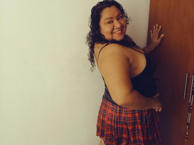 Curvy HiyunaMei with curly black hair, smiling, wearing a black tank top and red plaid skirt, standing against a wooden door, showing her big ass and small tits.