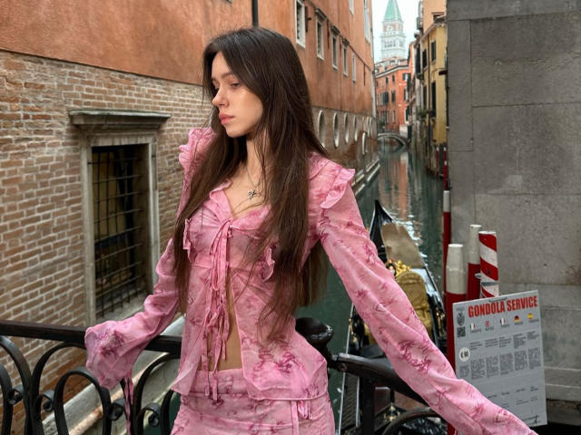 MoonSerenade with long brown hair, pink ruffled blouse, and matching pink skirt, standing by a canal, looking to the side, arms out, showing her small tits and slim ass.
