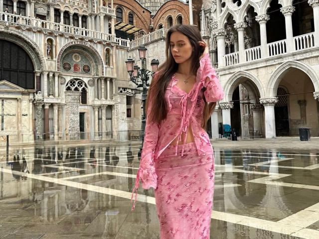 MoonSerenade with long brown hair, wearing a pink floral dress, stands in a wet courtyard, touching her hair, showing her small tits and flat ass.