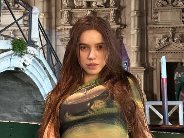 MoonSerenade with long brown hair, green and black patterned top, small tits, and a star necklace, standing in front of an ornate building with a blue arch and greenery.