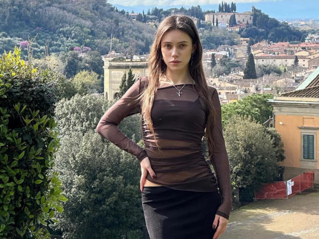 MoonSerenade with long brown hair, wearing a sheer black top, showing small tits, and a black skirt, standing with hands on hips, in a sunny, hilly, green neighborhood.
