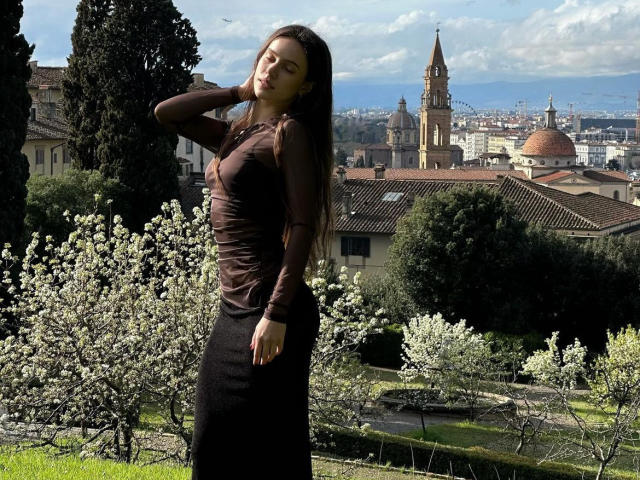 MoonSerenade stands in a sunlit garden, wearing a brown sheer top and black skirt, with long dark hair, her eyes closed, hand on neck, white blossoms in foreground.