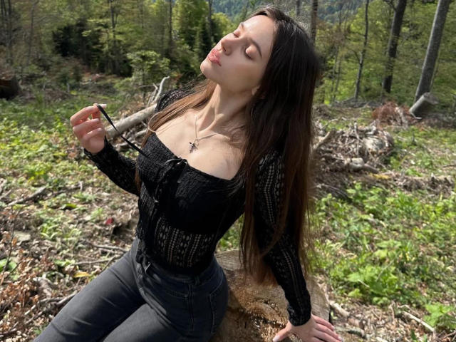 MoonSerenade with long brown hair, eyes closed, black off-shoulder top, holding a stick, sitting on a log in a forest, wearing black jeans, and a necklace.