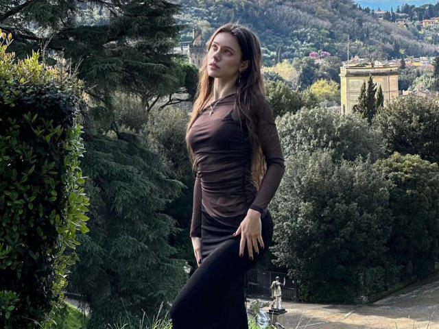 MoonSerenade stands with long brown hair, wearing a sheer brown top and black pants, her hand on her hip, showing her tits slightly, in a lush green garden.