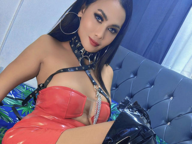 AddictiveMiss4u-hot in a red, shiny, latex dress with black leather straps and collar, blue eyes, red lipstick, hoop earrings, black gloves, and thigh-high boots, sitting on a blue bed.