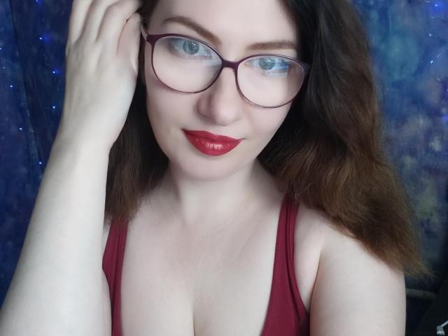 PaulinaSunny with red lipstick, glasses, and wavy brown hair, touching her head, showing her tits in a red tank top, blue eyes, fair skin.
