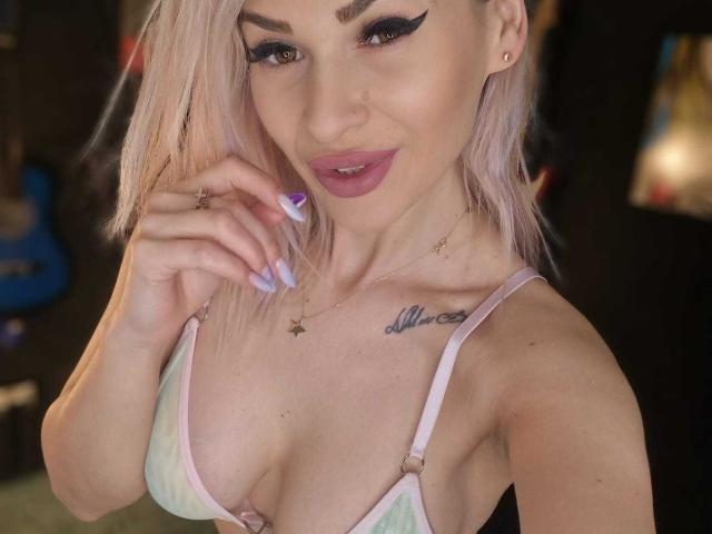 SelenaFendye con i capelli rosa, l'eyeliner scuro e il rossetto rosa, indossa un reggiseno pastello, mostrando il seno, un tatuaggio sopra il capezzolo sinistro e le unghie viola, sorridendo..
