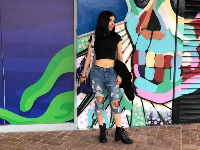 MileyWatson with black crop top, ripped blue jeans, and black boots, holding a black jacket, standing in front of colorful graffiti, smiling, with long black hair.