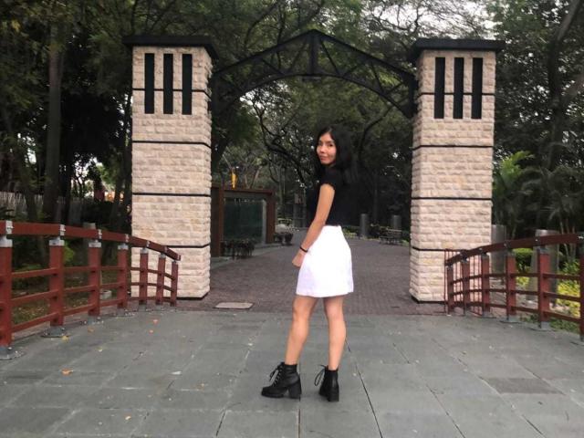 MileyWatson with black top, white skirt, black boots, standing in front of stone archway, looking back, medium tits, long dark hair, small ass, in a park setting.
