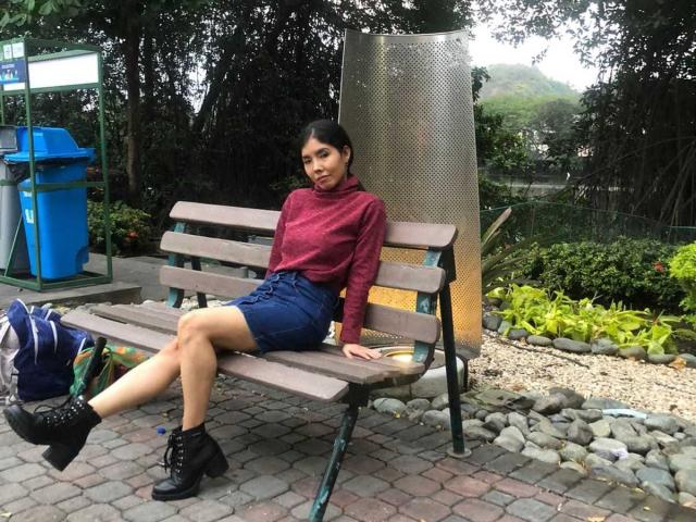 MileyWatson with small tits, dark hair, red sweater, blue shorts, black boots, sitting on wooden bench, legs crossed, backpack on ground, metal cylinder behind, greenery and rocks.