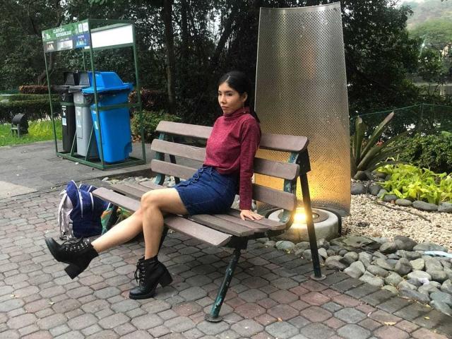 MileyWatson, sitting on a wooden bench, wearing a red sweater, blue shorts, black ankle boots, with her legs crossed, looking slightly to the side, in a park.