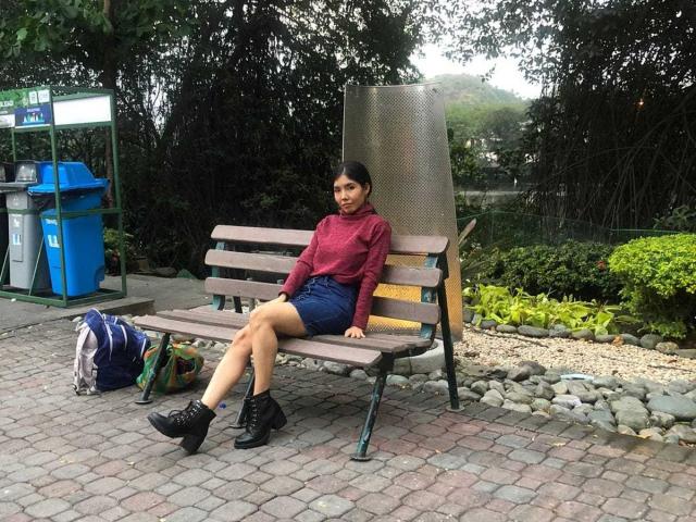 MileyWatson sits on a wooden bench, legs crossed, wearing a red sweater, blue shorts, and black boots, with a backpack on the ground, in a green park.