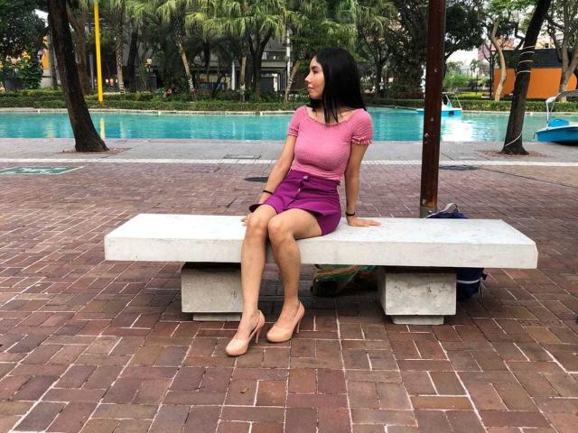 MileyWatson, with dark hair, sits on a white bench wearing a pink top and purple skirt, showing her ass, with tan heels, in a brick-paved, tree-lined area.