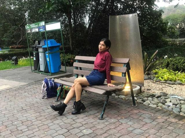 MileyWatson with black boots, red sweater, blue shorts, sitting on a bench, legs crossed, looking to the side, near a blue trash can, and a metal wall.