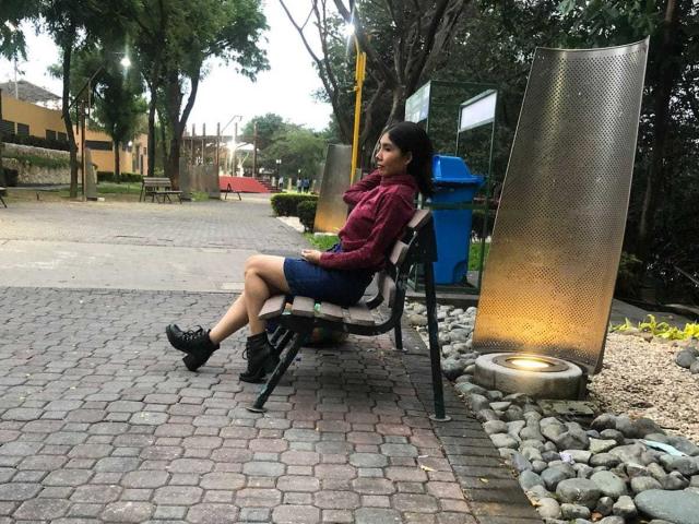 MileyWatson, with small tits, sits on a black chair, legs crossed, wearing a red shirt, blue skirt, black shoes, and black hair, in a park with a metal screen.