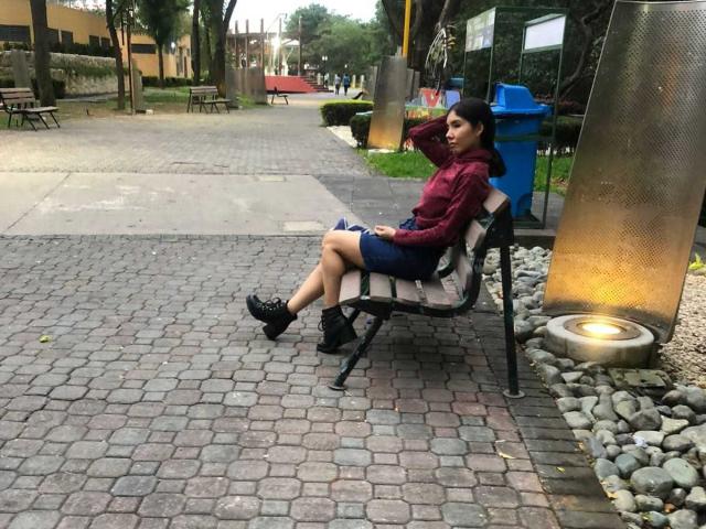 MileyWatson with dark hair, red hoodie, blue skirt, and black shoes, sitting on a bench, legs crossed, hands behind head, next to a metal lamp on a cobblestone path.