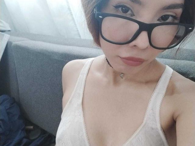 MileyWatson with glasses, wearing a white lace tank top, showing her small tits and a black choker with a tiny triangle pendant, looking at the camera.