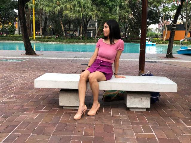 MileyWatson with black hair, pink top, and purple skirt, sitting on a white bench, legs crossed, wearing beige flats, looking to the side. Ass, pussy, tits visible.