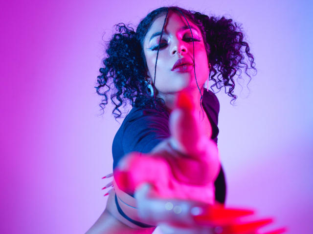 VioletHotX with curly black hair, black top, reaching forward with a red dildo in front, purple and pink neon light on her face, looking seductive, half-closed eyes.