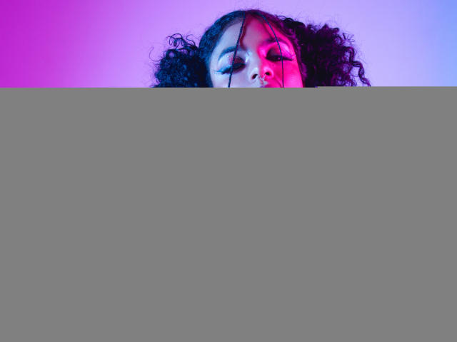 VioletHotX with curly black hair, reaching forward with a red dildo, wearing a black top, illuminated by purple and pink neon lights. Her face shows a slightly open mouth.