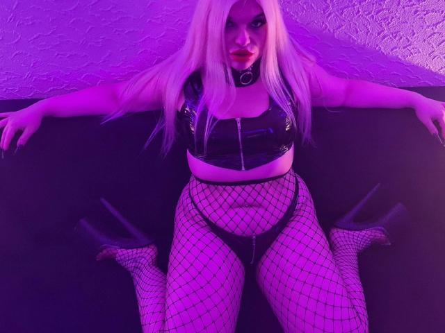 MariellaSun-hot with platinum blonde hair, black leather top, ring choker, fishnet pants, and high heels, sitting with arms spread, purple neon lighting, and red lipstick.