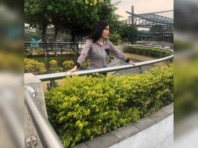 MileyWatson has black hair, wearing a grey button-up shirt, standing with hands on a metal railing, green bushes in front, and a bridge in the background.