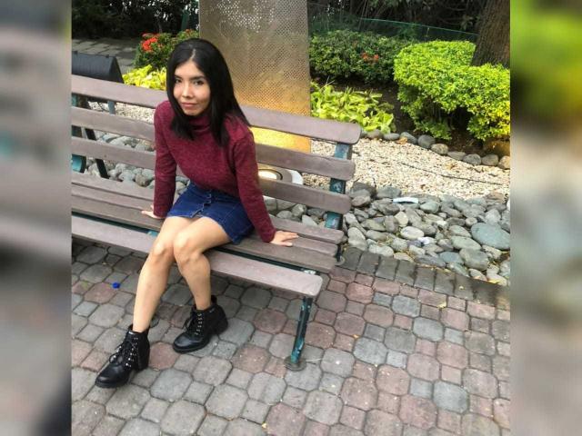 MileyWatson sitting on a wooden bench, wearing a red turtleneck, blue denim skirt, and black boots, with dark hair, looking at the camera, in a park.