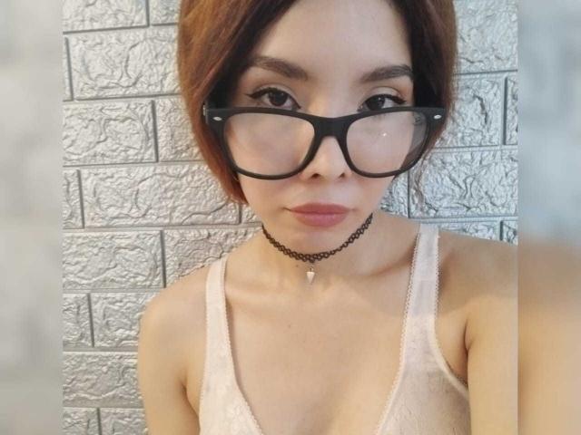 MileyWatson with glasses, brown hair, wearing a choker and white lace bra, showing her tits, standing against a textured brick wall.