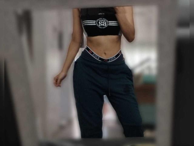 MileyWatson with a slim, toned midsection, wearing a black sports bra and dark blue pants, her ass slightly highlighted by the waistband. Her hand is partially in frame.