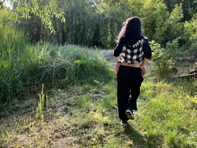 JennaEasy with curly black hair, wearing a black and white checkered shirt and black pants, walking away on a grassy path, sunlight filtering through greenery.