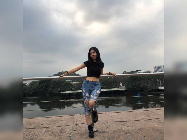 MileyWatson with dark hair, black crop top, and ripped blue jeans, standing on a stone bridge, arms out, looking at camera, cloudy sky, water reflection, and city buildings.
