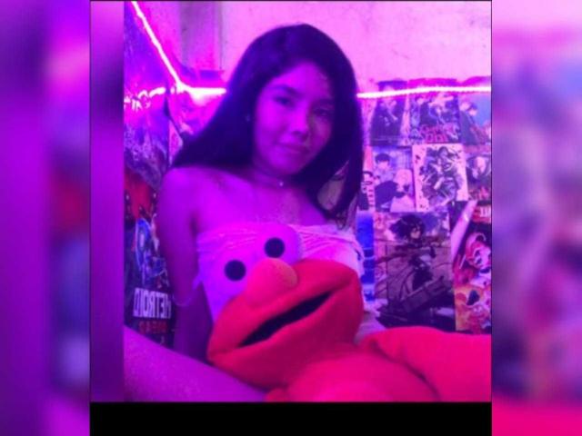 MileyWatson with long dark hair, wearing a white strapless top, sitting with a red Elmo plushie, in purple-lit room, surrounded by colorful posters.
