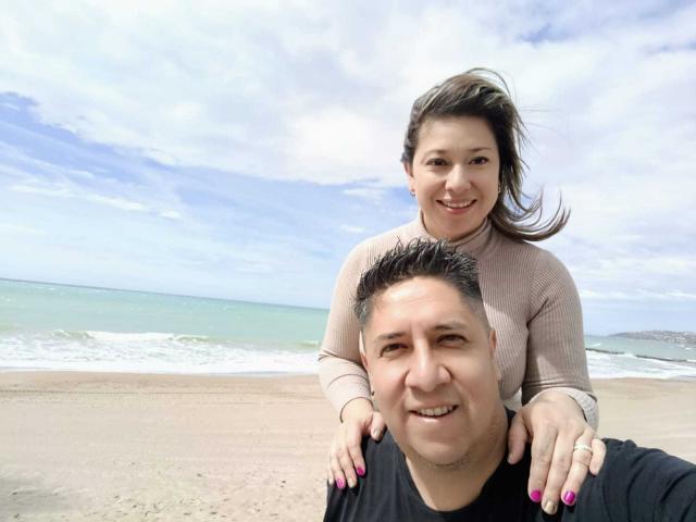 ThiagoEva with tits up, smiling, brown hair, tan sweater, hands on black-shirted ThiagoEva's head, beach, cloudy sky, ocean, ThiagoEva's pink nails, ThiagoEva's short black hair.
