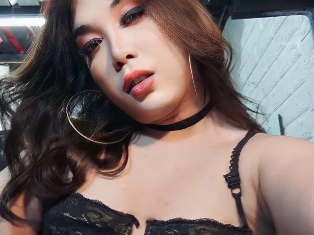 SuperHotVenna with wavy brown hair, wearing black lace bra, choker, and hoop earrings, has red lipstick and looks down with slightly parted lips. Boobs visible in the shot.