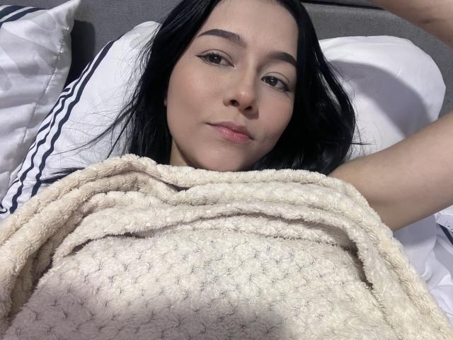 DeisyWest, lying in bed, covered with a beige, textured blanket, has black hair, light skin, and is looking at the camera with a slight smile, arms raised.