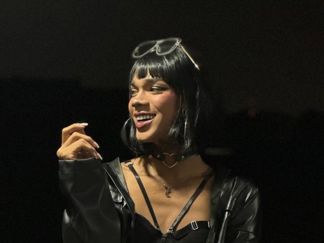 NaomiSteward with short black hair, wearing a black choker, leather jacket, and glasses on her head, smiling with her hand up, showing her teeth and ring. Dark background.