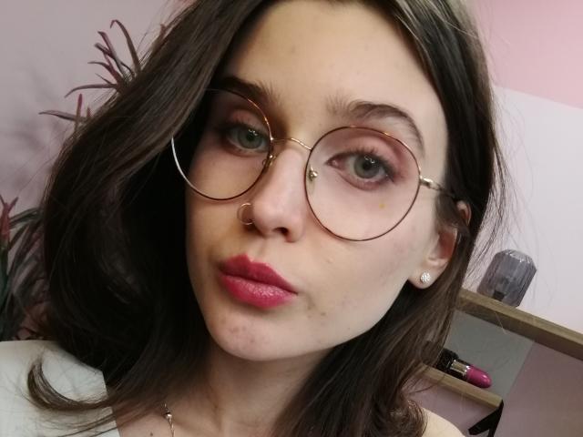 SarahMiss with round glasses, pink lipstick, nose ring, and earrings, has pale skin, green eyes, and brown wavy hair, showing a slight pout. Her tits are not visible.