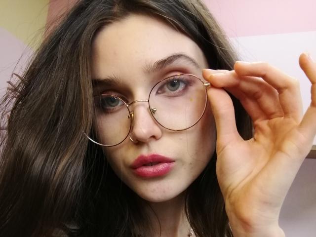SarahMiss with long brown hair, wearing gold-rimmed glasses, pink lipstick, and a nose ring, adjusting her glasses with her right hand, has a serious expression.