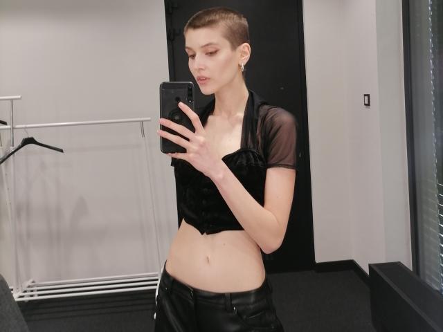 SarahMiss with a shaved head, wearing a black sheer top and leather pants, taking a mirror selfie with her phone. She has small tits and a slim, pale body.