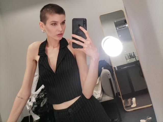 SarahMiss with a shaved head, wearing a black pinstripe halter top and matching skirt, holding a phone in a mirror selfie, showing a tattoo on her right arm.