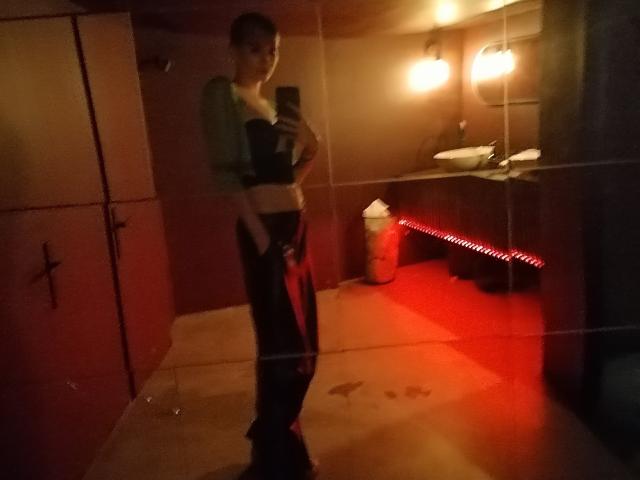 SarahMiss with short brown hair, green top, black bra, and loose pants, standing in dimly lit room, taking mirror selfie, red lighting on floor, holding phone.