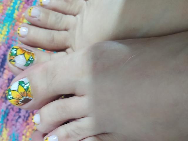 DalilaSuperHots Füße mit weißem Nagellack und gelben Sonnenblumen-Nagelmotiven an den Zehen, wobei ein rosa Hautton und ein farbenfroher, unscharfer Hintergrund zu sehen sind..