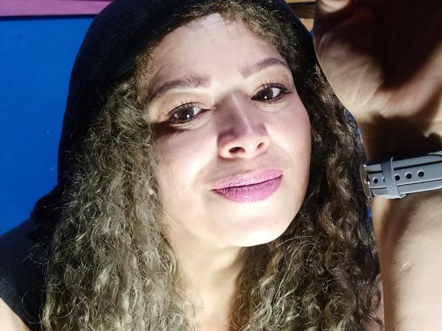 DalilaSuperHot with curly brown hair, wearing a black hood, pink lipstick, and a gray wristband, smiling softly at the camera. Her cute ass is partially visible on the right.