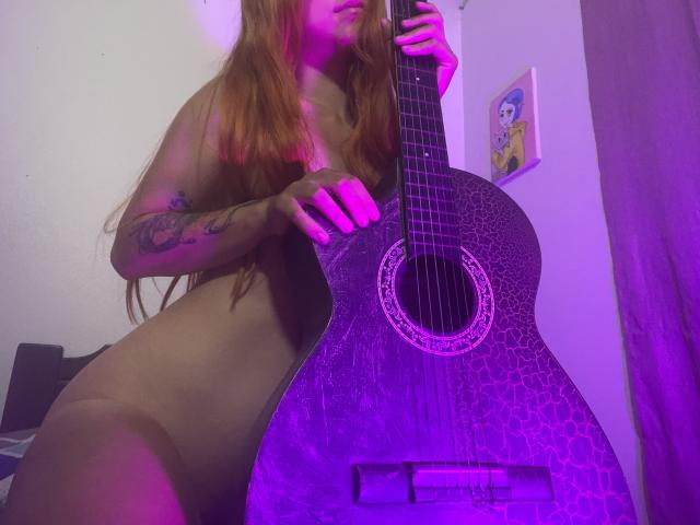 JuliaGarciax, naked with long red hair, holding a purple-accented guitar, tattoos on left arm, purple lighting, her tits partially visible, her hand on the guitar's neck.