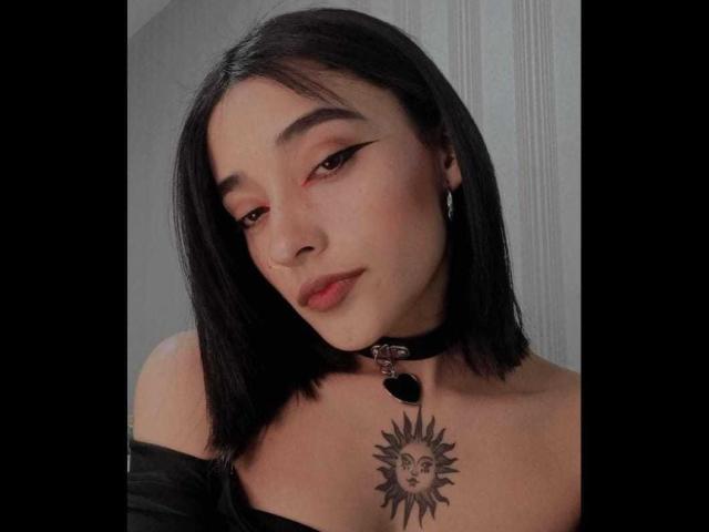 SalomeMille with black shoulder-length hair, wearing a black choker with a sun tattoo on her chest, eyeliner, and red eye makeup, looking slightly down.
