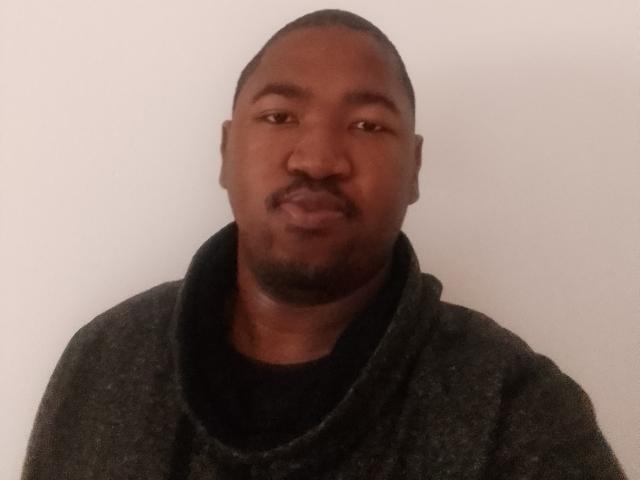 BigDickCya261, a dark-skinned dude with a goatee, short hair, and a slight smile, wearing a gray, cowl-neck sweater, looking straight at the camera.