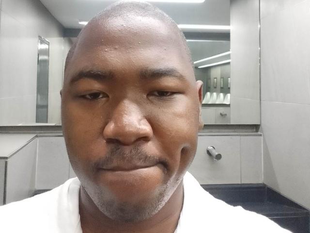 BigDickCya261 has a bald head, dark brown skin, a small mustache, and a goatee, wearing a white shirt, with a slight smirk in a bathroom selfie.
